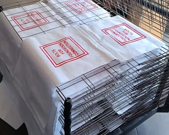 Tek renk serigrafi baskılı etiketler kurutma rafında — kırmızı mürekkeple basılmış uyarı metni, tel raflarda istiflenmiş | Divan Etiket Ankara